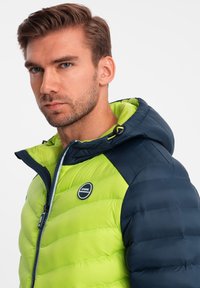 Vihreä ja laivastonsininen toppatakki hupulla, jossa on vetoketjukiinnitys, vaakasuora tikkaus ja pyöreä logo rinnassa.