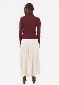 Maglione aderente bordeaux a collo alto con maniche lunghe, abbinato a pantaloni crema a gamba larga, che mostra una texture liscia e un contrasto di colori neutri.