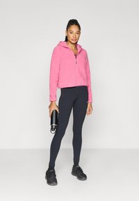 Polarowy bluza z kapturem i zamkiem błyskawicznym w jasnoróżowym kolorze, połączona z czarnymi legginsami. Modelka trzyma czarną butelkę na wodę i ma na sobie czarne buty sportowe.