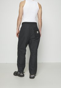 Czarne spodnie robocze o luźnym kroju, z elastycznym paskiem w talii oraz tylną kieszenią z naszywką z logo. Noszone z czarnymi klapkami.