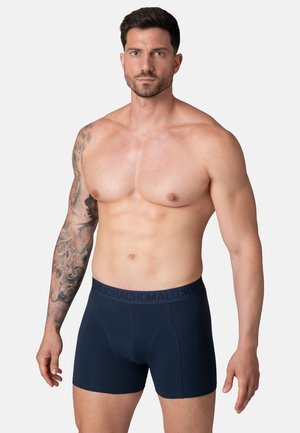 Spierkrachtige man met een getatoeëerde linkerarm, gekleed in marineblauwe boxershorts met een merknaam op de tailleband, staand tegen een effen lichte achtergrond.