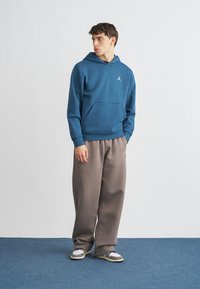 Μπλε φούτερ με κουκούλα και μπροστινή τσέπη, με λευκό λογότυπο, σε συνδυασμό με γκρι φόρμα. Το μοντέλο φοράει γκρι και άσπρα αθλητικά παπούτσια. Ομαλή υφή υφάσματος.