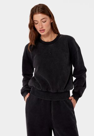 Kobieta nosząca luźną czarną bluzę i dopasowane spodnie, z rękami w kieszeniach, patrząca w dół na jednolitym jasnym tle.