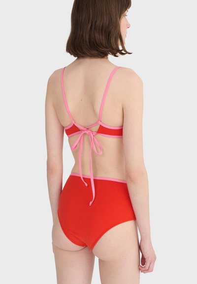 Femme aux cheveux bruns courts portant un bikini rouge avec un bord rose, vue de dos sur un fond gris clair uni.