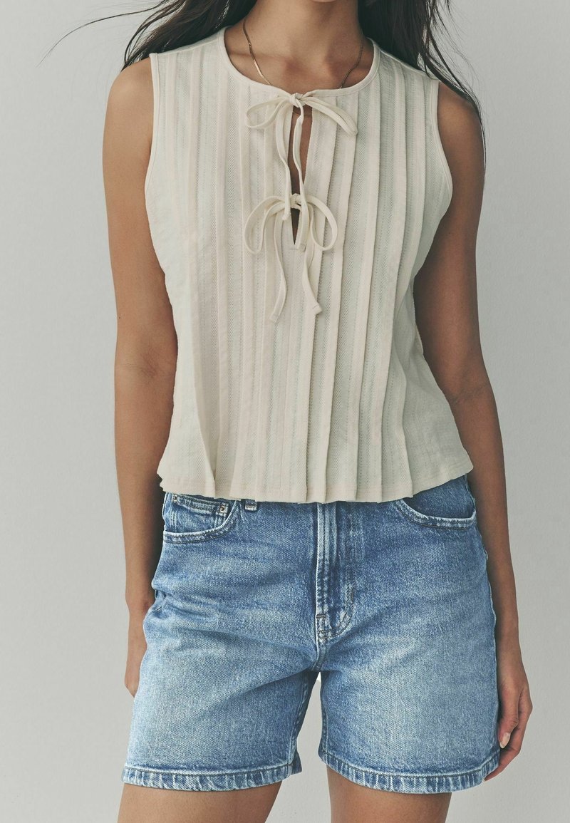 Femme portant un haut crème sans manches en maille avec deux liens à l'avant et un short en denim bleu taille haute, debout devant un fond uni.