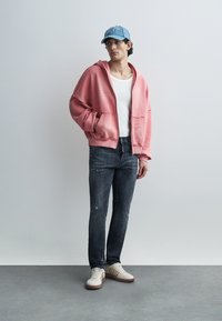 Ροζ φαρδιά φούτερ με τσέπες μπροστά, φορεμένο πάνω από λευκό αμάνικο τοπ, σετ με σκισμένα σκούρα τζιν και μπεζ αθλητικά παπούτσια. Ανοιχτό μπλε καπέλο.