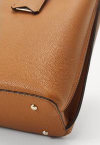 Bruine leren tas met een specifieke textuur, voorzien van gouden hardwareaccenten en contrasterende zwarte piping langs de randen.