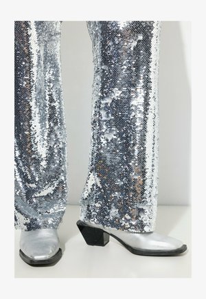 Pantaloni a zampa d'elefante argentati con paillettes abbinati a stivali alla caviglia in pelle argentata con tacco angolare nero. La texture lucida aumenta l'appeal visivo.