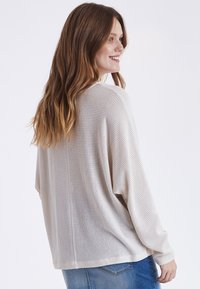 Leichter, offen gestrickter cremefarbiger Pullover mit überschnittenen Schultern und langen Ärmeln, kombiniert mit blauen Jeans. Sanfte Textur, lockere Passform.