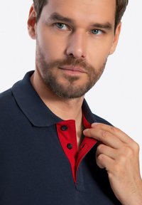 Námořnická polo košile s červeným límečkem, která má dvoubarevný knoflíkový zapínání. Hladká textura, těsný střih a krátké rukávy jsou viditelné.