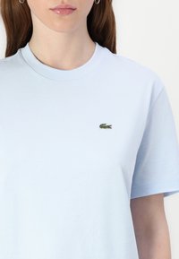 Camiseta de algodón de color azul claro con cuello redondo y mangas cortas. Tiene un pequeño logo de cocodrilo bordado en el lado izquierdo del pecho.