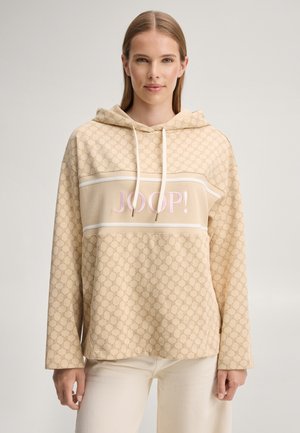 Femme aux cheveux lisses châtain clair porte un sweat à capuche beige à motifs avec "JOOP!" en rose et un pantalon blanc, debout devant un fond uni.