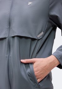 Χέρι στην τσέπη ενός γκρι μπουφάν με φερμουάρ και το λογότυπο της Fila στο στήθος και ανακλαστική ταινία κοντά στην τσέπη.