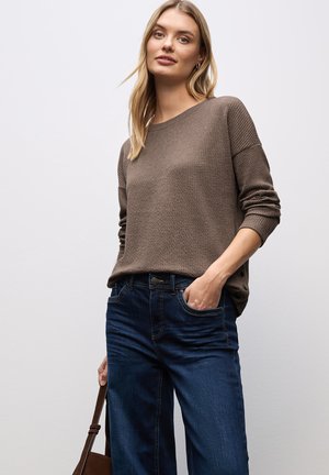 Suéter marrón de punto con un patrón texturizado, combinado con jeans azul oscuro que tienen bolsillos y cierre de botón. Bolso marrón colgado sobre el hombro.