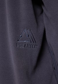 Tamsiai pilkas švelnus audinys su tekstūruota paviršiumi. Išsiuvinėtas "POLARTEC" logotipas su trikampiu dizainu ir rodyklėmis kontrastingos tamsiai mėlynos spalvos.