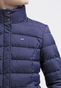 Navy modrá prošívaná bunda s vysokým límcem, zapínáním na cvoky a malým obdélníkovým logem na hrudi, kterou má na sobě osoba otočená vlevo.