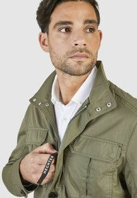 Veste vert olive avec fermeture éclair et bouton, poche poitrine et tissu texturé. Équipée d'un col montant et d'une chemise blanche en contraste en dessous.