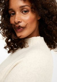 Naheaufnahme einer Person mit lockigem braunem Haar, die einen cremefarbenen Pullover mit hohem Kragen trägt. Der Pullover hat eine weiche Textur und einen gerippten Kragen.