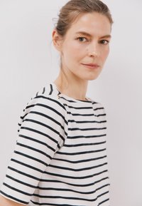 Proužkované tričko v bílé a námořnické barvě, s krátkými rukávy a kulatým výstřihem. Obsahuje zlatý akcent poblíž ramenního švu. Hladká textura látky.