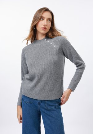 Grå strikkegenser med høy hals, ribbestruktur og dekorative knapper på skulderen. Kombinert med blå vide jeans.