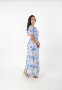 Robe maxi à motif floral bleu clair, en tissu transparent avec des manches bouffantes, taille cintrée, jupe fluide, portée avec des sandales à talons transparents.