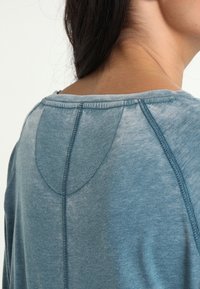 Miętowozielony, miękki materiałowy top z zaokrąglonym dekoltem i widocznymi szwami, z zakrzywionym tylnym karczkiem. Subtelna tekstura i swobodny design.