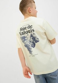Kreminės spalvos marškinėliai su vynuogių iliustracija ir užrašu "Rue de Valouis" ant nugaros; trumpomis rankovėmis ir laisvu pavyzdžiu.