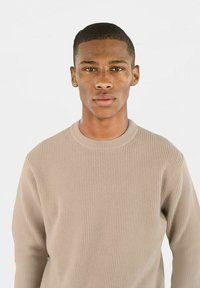 Beige ribbestrikket genser med rund hals og lange ermer, med en glatt tekstur og tettsittende silhuett. Ingen mønstre eller aksenter.