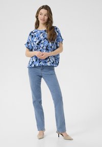 Blaue abstrakte gemusterte Bluse mit lockerer Passform, kurzen Ärmeln und rundem Ausschnitt, kombiniert mit geraden, hellen Jeans und beigen Absätzen.