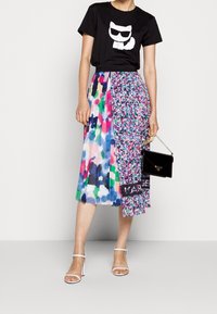 Camiseta negra con gráfico de gato blanco, combinada con una falda de longitud hasta la rodilla de colores multifacéticos que presenta un patrón vibrante. Sandalias de tacón bloque blanco y bolso negro.