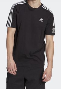 Muž v černém tričku Adidas s bílými třemi pruhy na rukávech a logem Adidas na hrudi a levém rukávu.