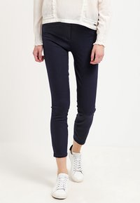 Pantalones de stretch en un azul marino oscuro con un diseño ajustado, textura suave y cintura de altura media, combinados con zapatillas blancas y una blusa clara.