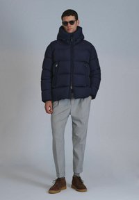 Marineblaue Steppjacke mit hohem Kragen und verstellbarem Kapuze, ausgestattet mit Reißverschlusstaschen. Kombiniert mit hellgrauen Hosen und braunen Stiefeln.
