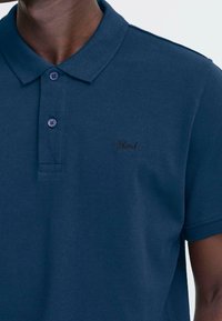 Mann iført en marineblå polo med knapper og en liten brodert "Blend"-logo på brystet.