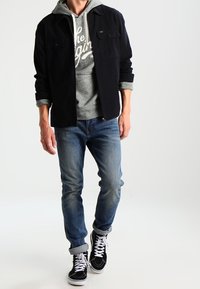 Giacca nera con bottoni sopra una felpa grigia con stampa bianca, jeans blu e sneaker alte nere con suole e lacci bianchi.