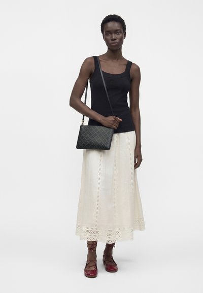 Femme portant un débardeur noir, une longue jupe crème avec des détails en dentelle, des chaussures rouges à lacets et un sac bandoulière noir avec un motif en losange.