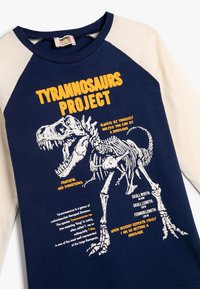 Niebieska i kremowa koszulka raglanowa z grafiką dinozaura, żółtym tekstem i szczegółami na temat anatomii Tyranozaura. Wykonana z bawełny.