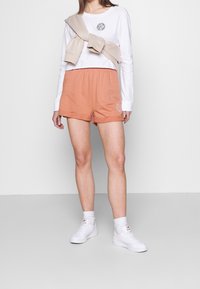 Vit långärmad skjorta med en logotyp, beige tröja knuten runt halsen och orangea shorts med sidobroderi; bärs med vita sneakers och strumpor.