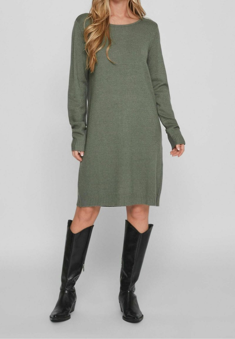Femme portant une robe en maille vert olive mi-longue à manches longues et des bottes en cuir noires montant jusqu'aux genoux, debout sur un fond gris.
