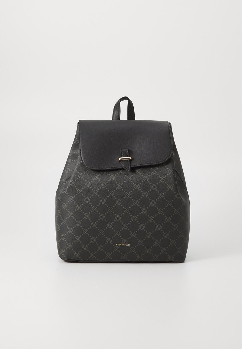 Sac à dos noir avec rabat et fermeture à boucle, orné de petits logos, avec une poignée supérieure sur un fond clair uni.