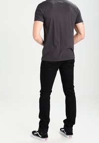 Man som står med ryggen mot kameran, iklädd en mörkgrå t-shirt, svarta slim jeans och svart-vita Vans sneakers mot en vit bakgrund.