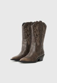 Botas de vaquero de cuero marrón con punta afilada, costuras decorativas y tachuelas de acento, con un tacón bajo apilado y tiradores laterales.