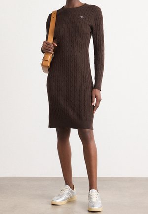 Osoba ubrana w brązową, długosleeową sukienkę z dzianiny warkoczowej, srebrne trampki, trzymająca beżową torbę na ramię na jednolitym tle.