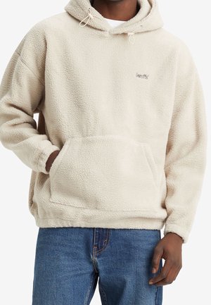 Sudadera de felpa beige con un bolsillo tipo canguro, capucha ajustable con cordón y pequeño logo bordado en el lado izquierdo del pecho. Textura suave.
