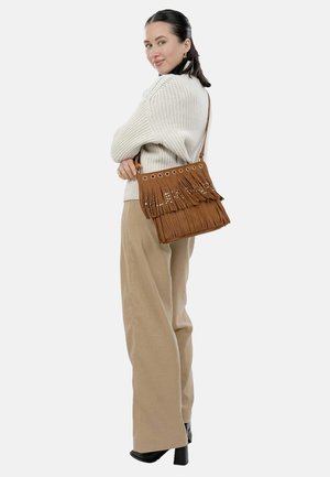 Braune Handtasche mit Fransen und Ösenakzenten, rechteckige Form, weiche Textur. Im Crossbody-Stil getragen mit einem leichten Pullover und beigen Hosen.