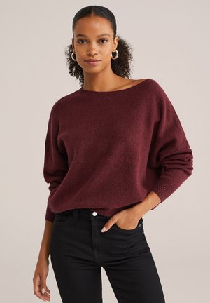Pull en tricot oversize bordeaux avec un décolleté large, des poignets côtelés et une texture douce, associé à un jean noir ajusté.