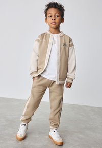 Veste polaire bicolore beige avec boutons-pression, t-shirt graphique blanc, joggeurs beige assortis et baskets blanches avec semelles en gomme.