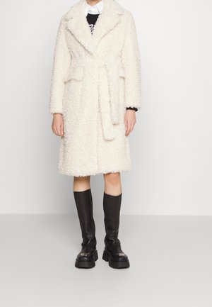 Manteau ivoire et duveteux avec une ceinture à nouer, grand col et poches latérales, associé à des bottes noires montantes avec des semelles chunky.