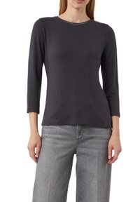 Dunkelgraues Langarmshirt mit rundem Ausschnitt, aus weichem Baumwollmaterial und einem dezenten Metallakzent am Kragen. Kombiniert mit hellgrauen Hosen.