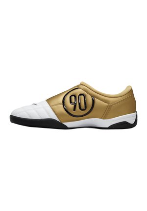 Niskoprofilowe halowe buty piłkarskie w kolorze złotym i białym z czarną podeszwą i logo „90” z boku.